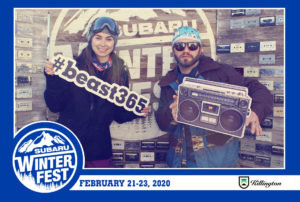 A man and a woman holding Subaru signs posing in the branded backdrop photo booth with a Subaru Winter Fest frame