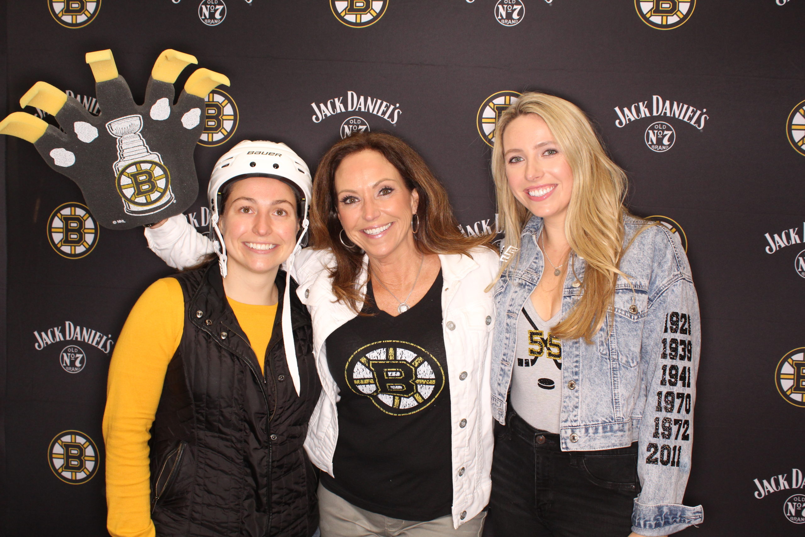 Three women wearing Bruins props posing for a photo in the Jack Daniel's/Bruins branded backdrop photo booth
