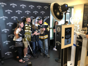 Overview of a group wearing Bruins gear pose for a picture in the Jack Daniel's/Bruins branded backdrop photo booth