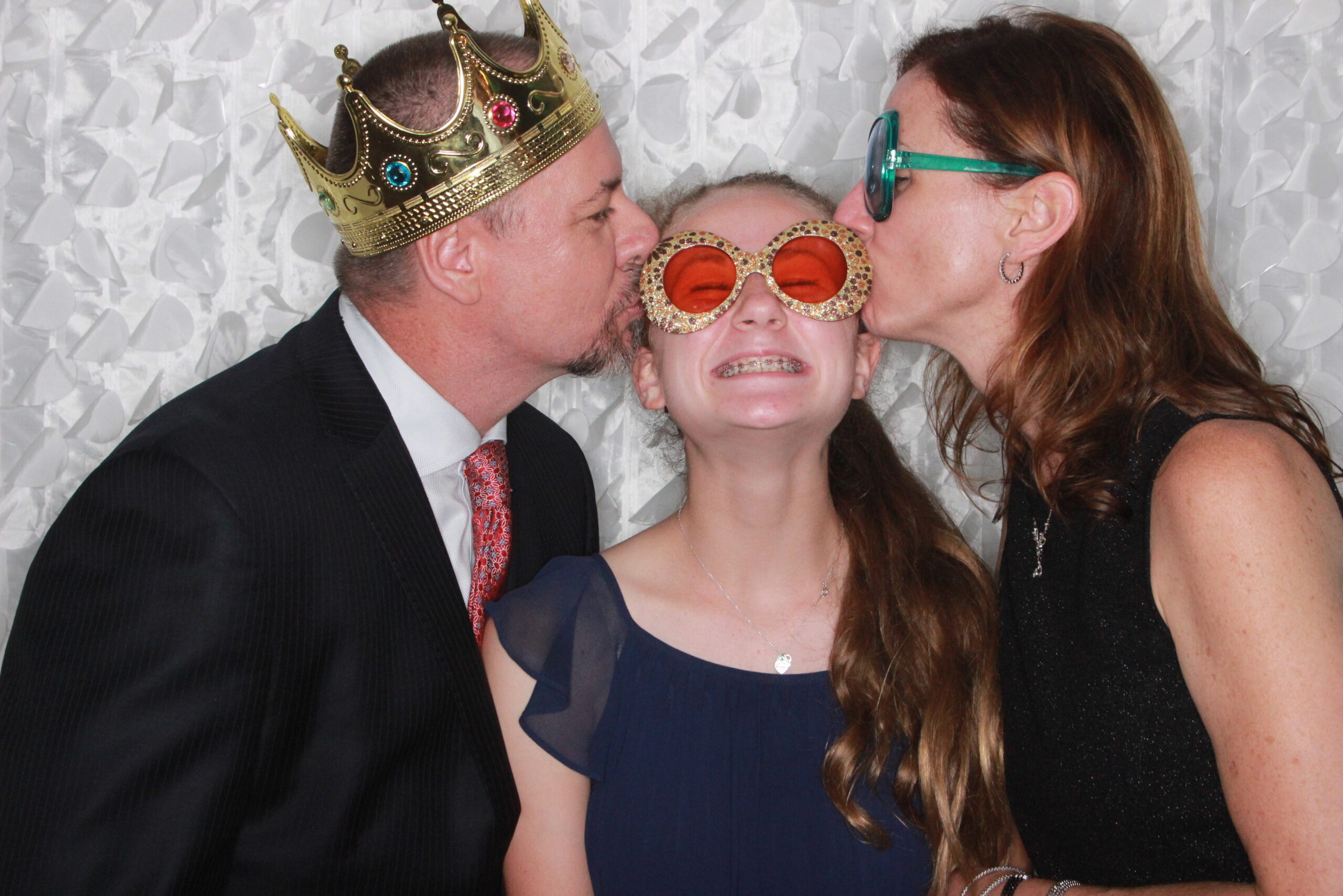 parents kissing their daughter on both sides of her head while wearing photo booth props
