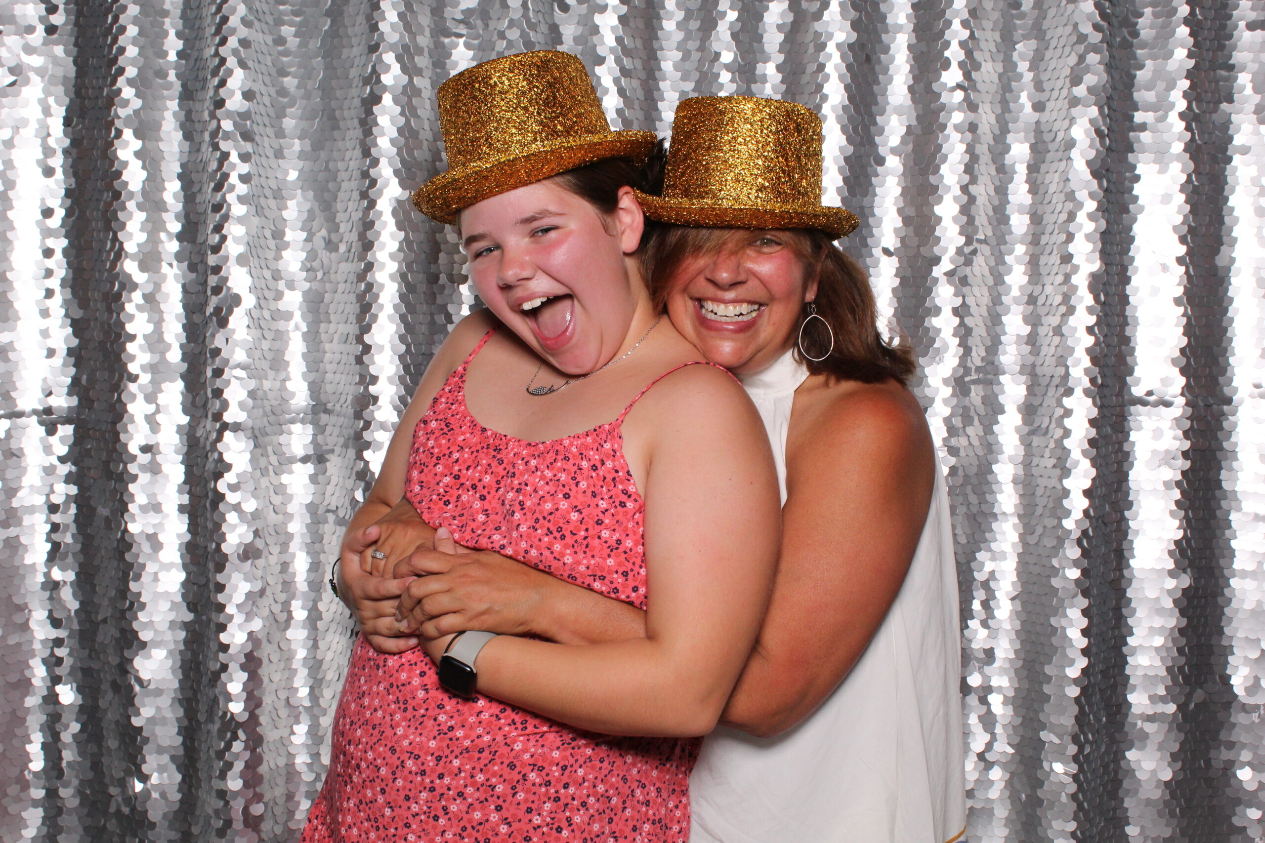 two women wearing sparkly gold top hats hugging and laughing while posing for the camera in a silver background photo booth