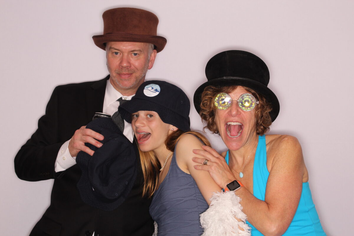 a girl and her parents pose for a silly picture in a white background photo booth while wearing photo booth props
